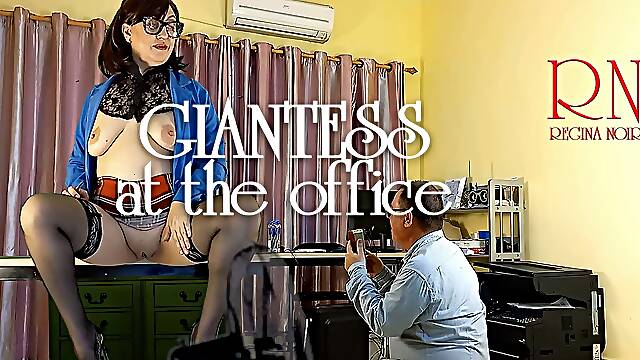 A giant secretary sits on an office desk. The manager guy is very surprised at her height.